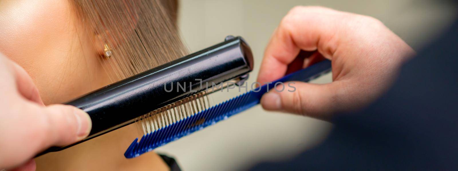 A hairdresser is straightening the hair of the young brunette woman in a beauty salon. by okskukuruza