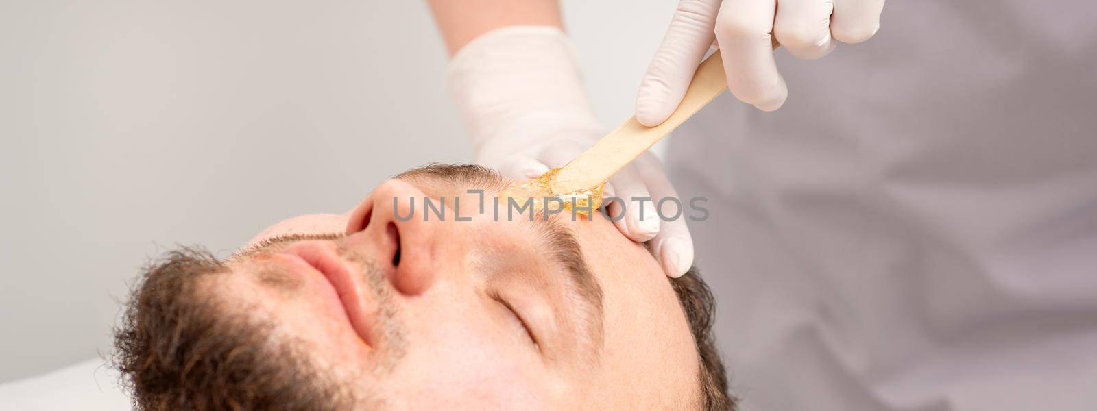 Beautician applying wax paste between eyebrows during the procedure of waxing in the beauty salon