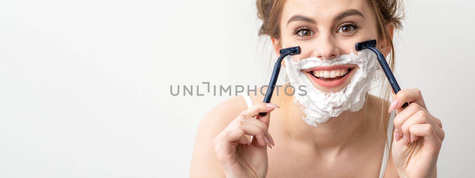 Beautiful young caucasian woman shaving her face by razor on white background. Pretty smiling woman with shaving foam and razor on her face