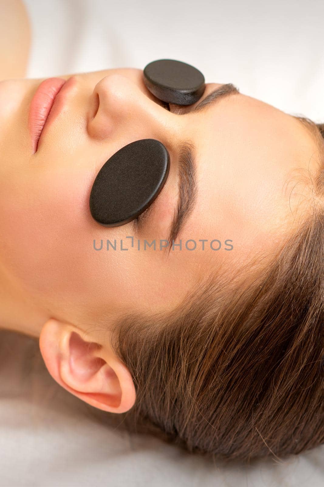 Black massage stones lying on the eyes of the young caucasian woman. Facial massage in a spa