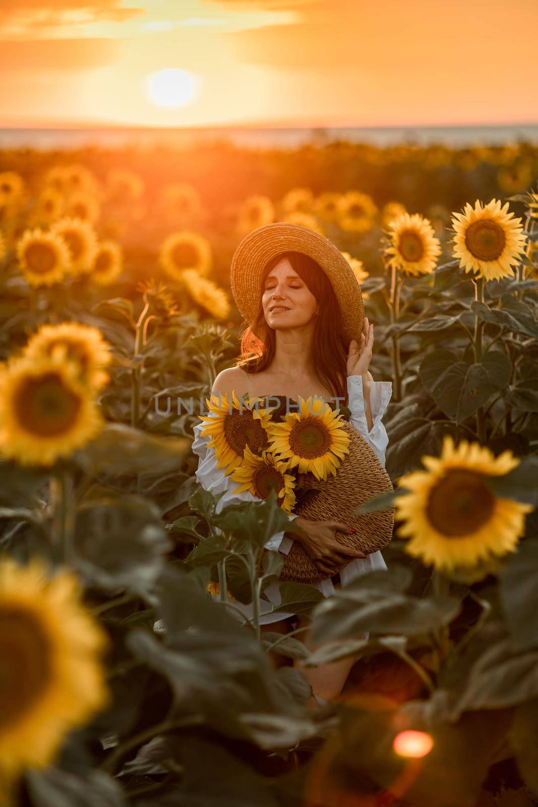 A girl in a hat on a beautiful field of sunflowers against the sky in the evening light of a summer sunset. Sunbeams through the flower field. Natural background