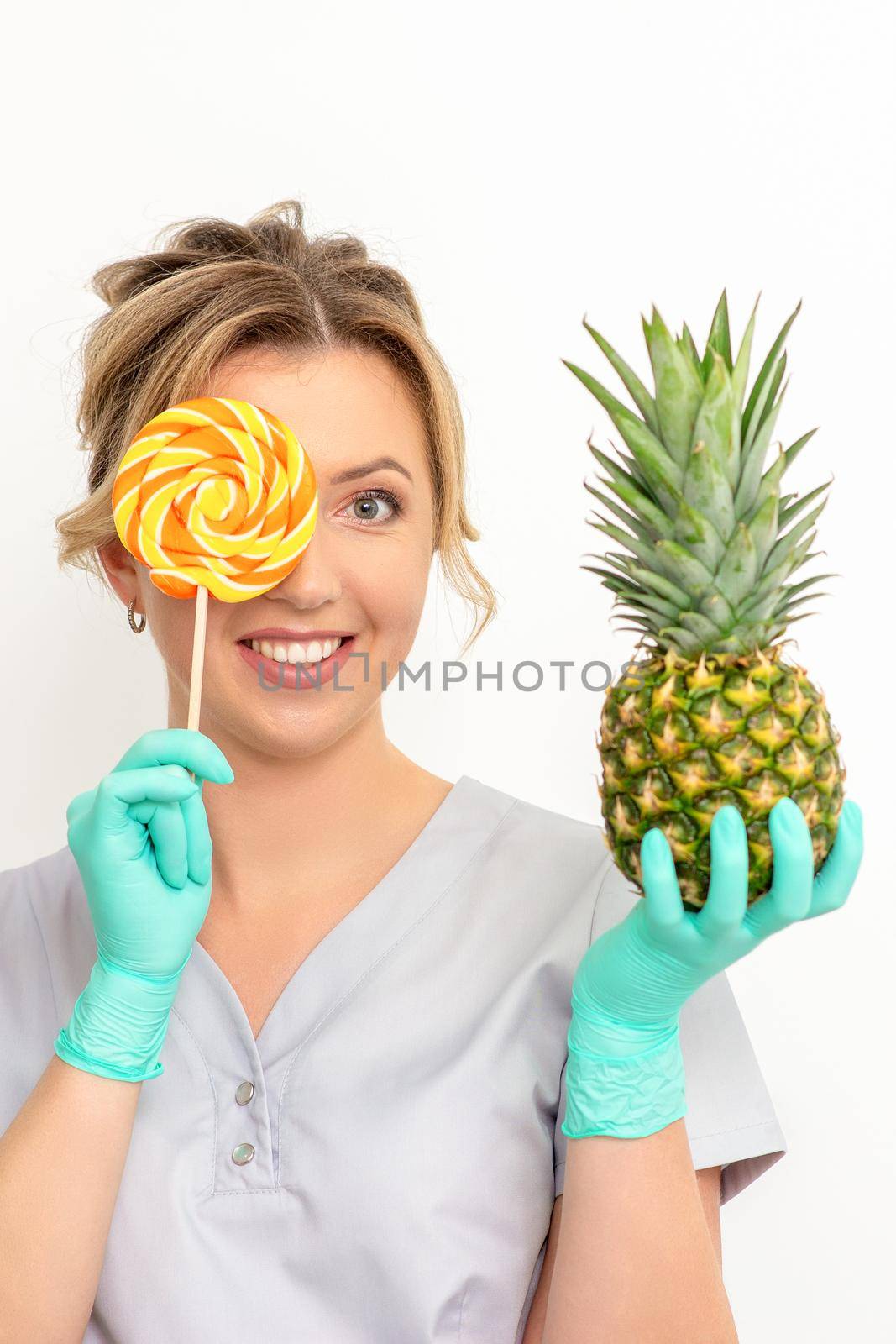 Beautiful smiling beautician holding fresh pineapple and cover her eye with lollipop over white background. Skincare cleansing eco organic