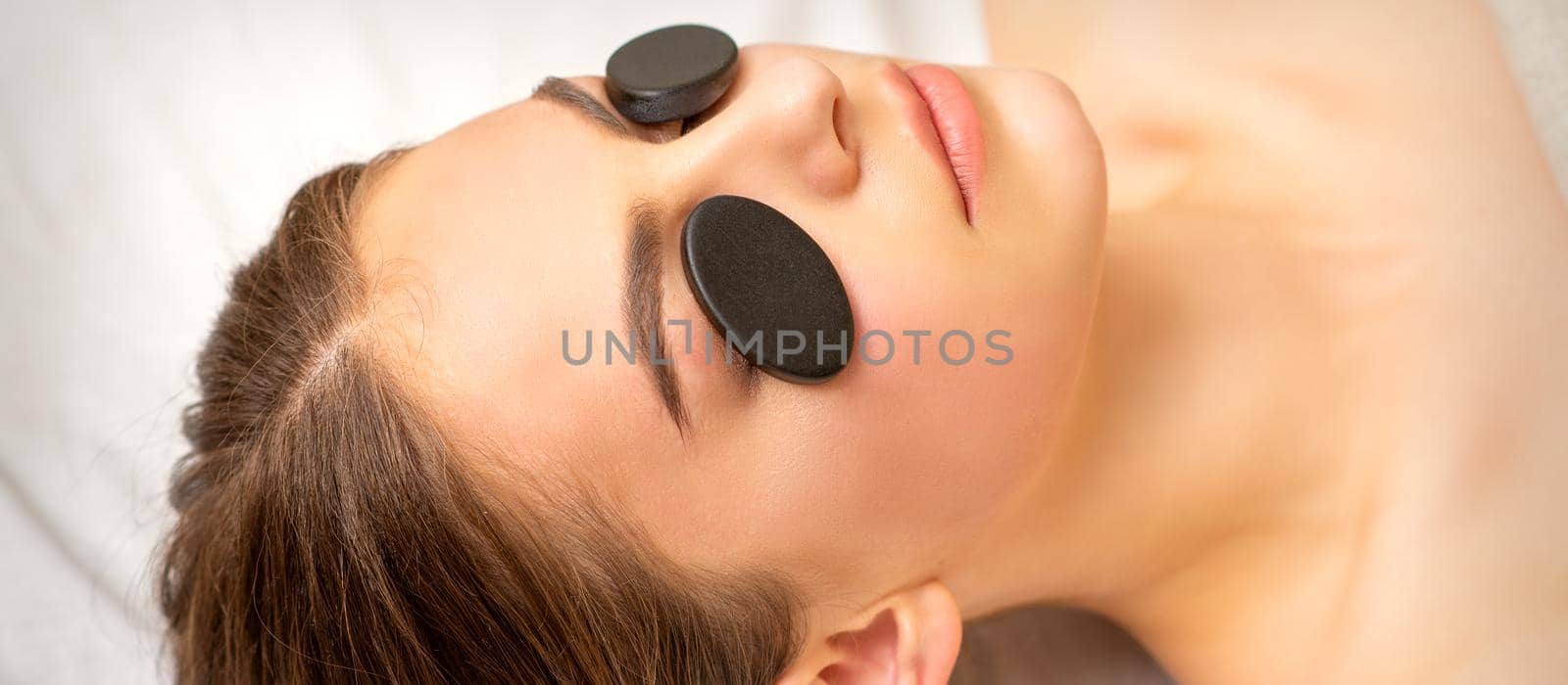 Black massage stones lying on the eyes of the young caucasian woman. Facial massage in a spa