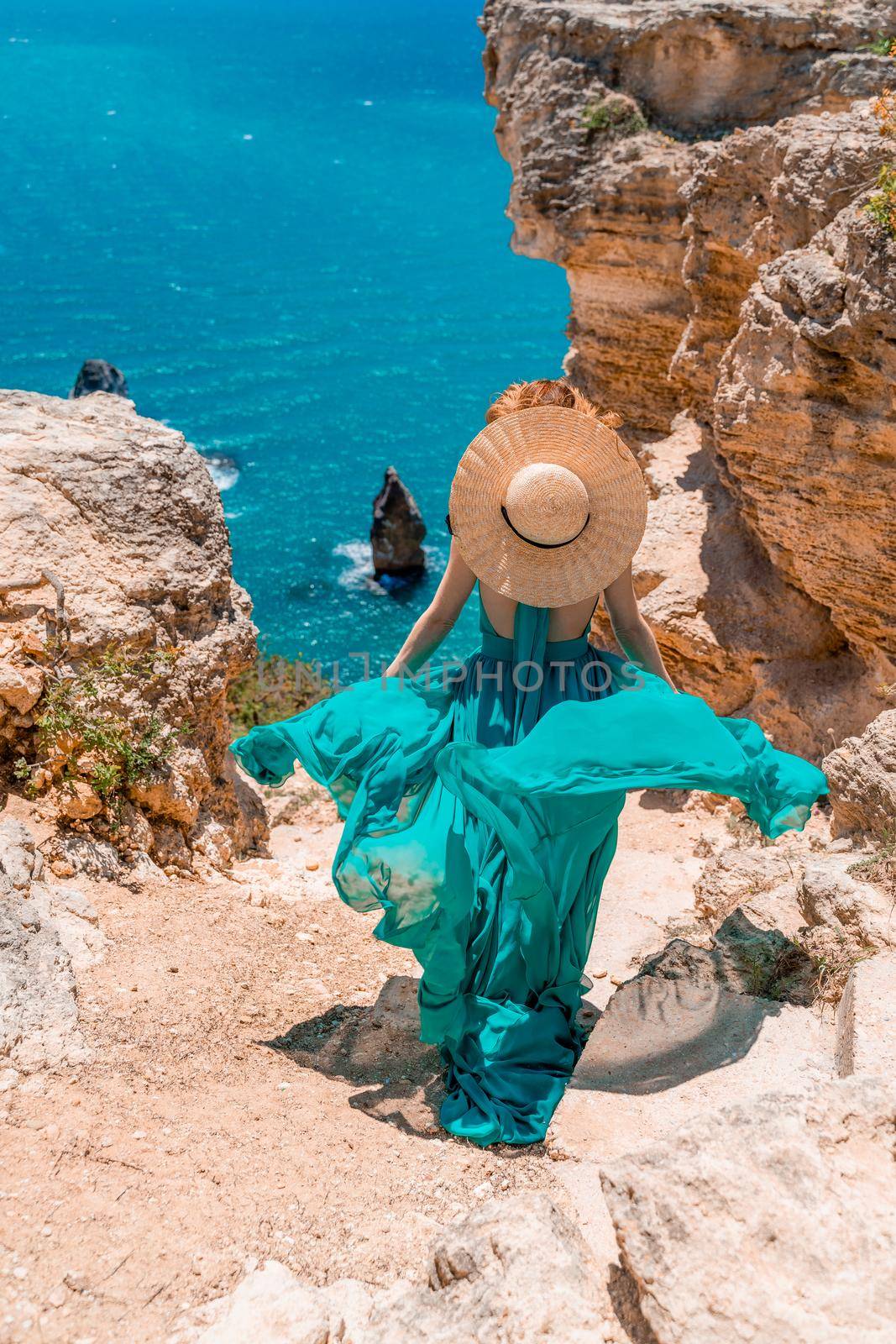 A girl with loose hair in a long mint dress descends the stairs between the yellow rocks overlooking the sea. A rock can be seen in the sea. Sunny path on the sea from the rising sun by Matiunina