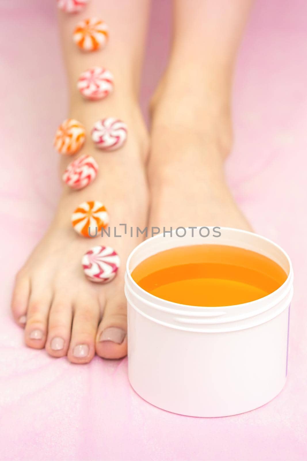 Round candies lying in a row on female feet with white jar with sugar paste on pink background with copy space, depilation concept. by okskukuruza