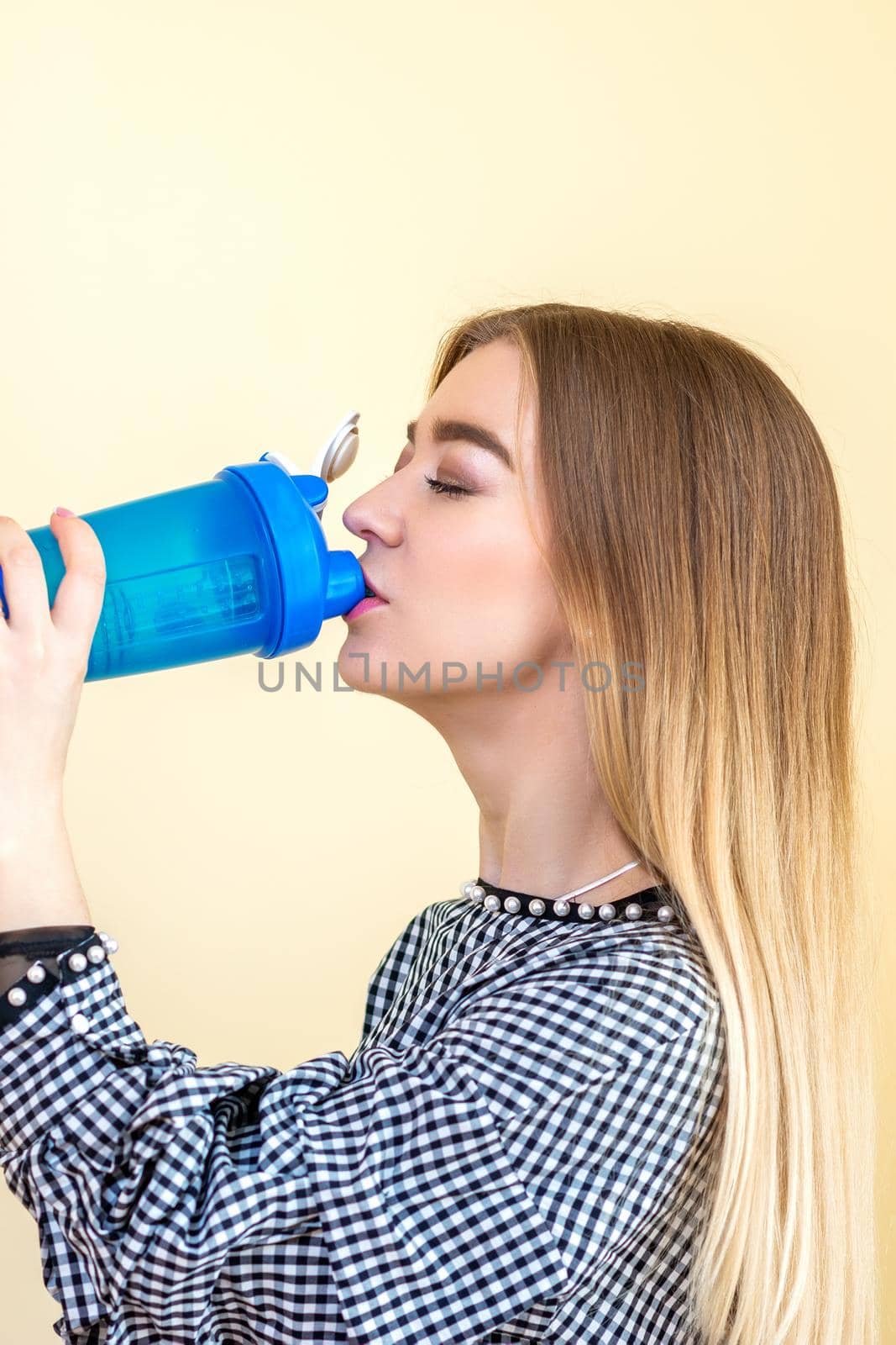 Thirsty woman with blue plastic bottle against a light background. A beautiful young caucasian businesswoman in a blouse drinking water on yellow