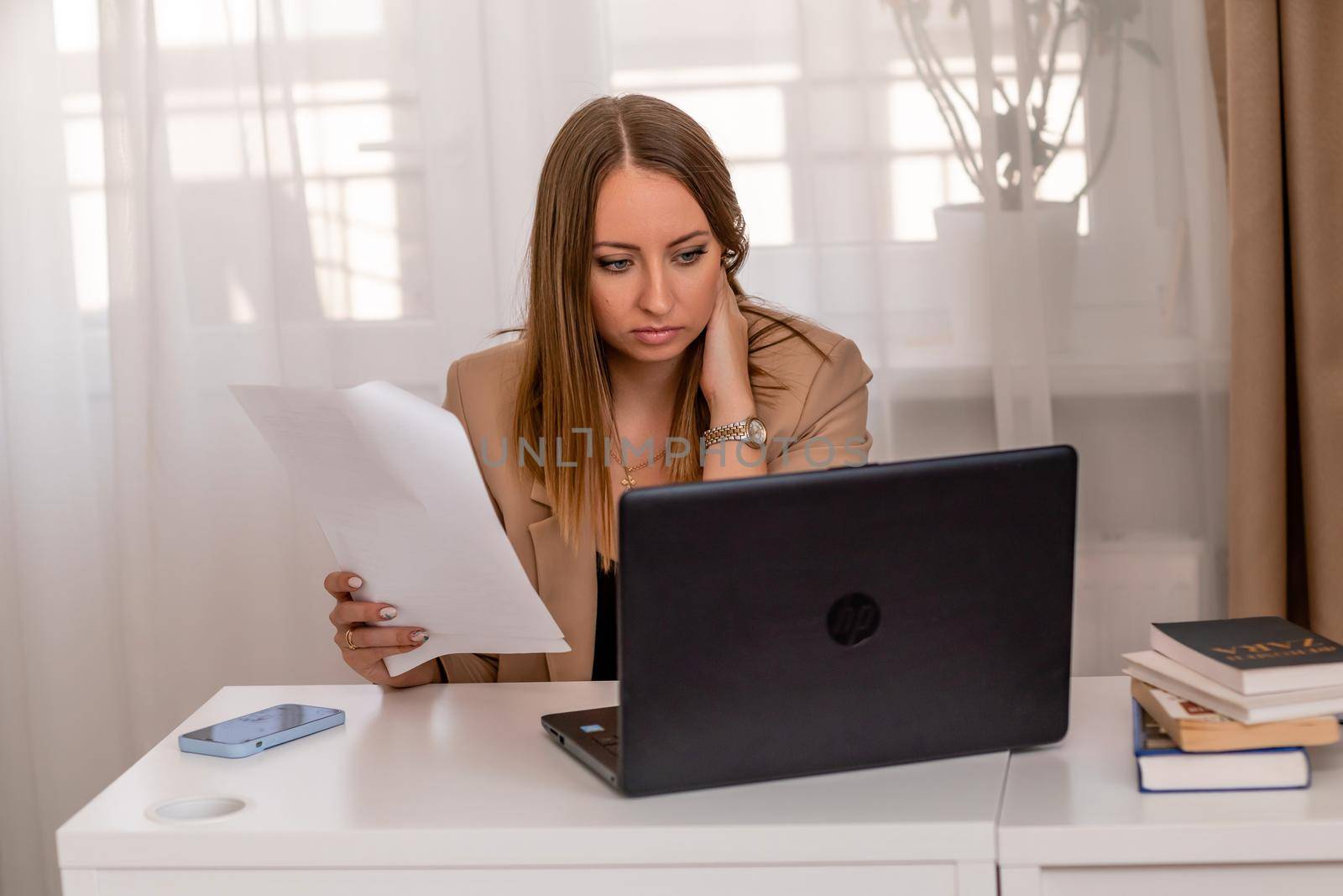 European professional woman sitting with laptop at home office desk, positive woman studying while working on PC. She is wearing a beige jacket and jeans. by Matiunina
