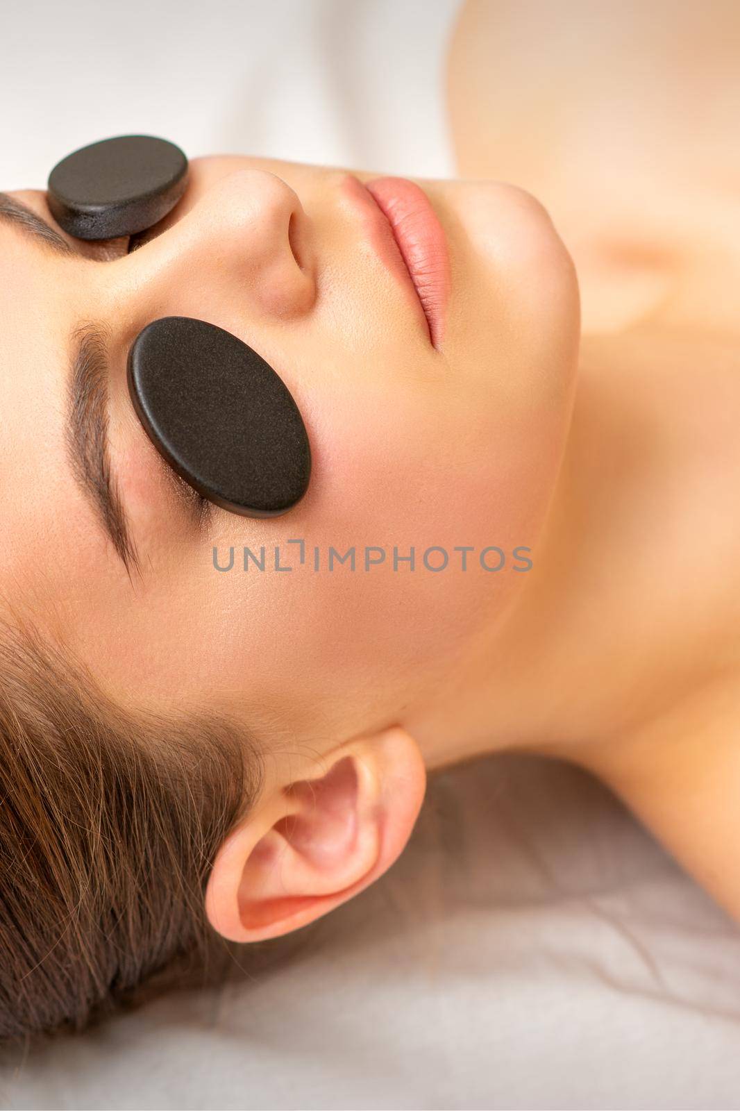 Black massage stones lying on the eyes of the young caucasian woman. Facial massage in a spa