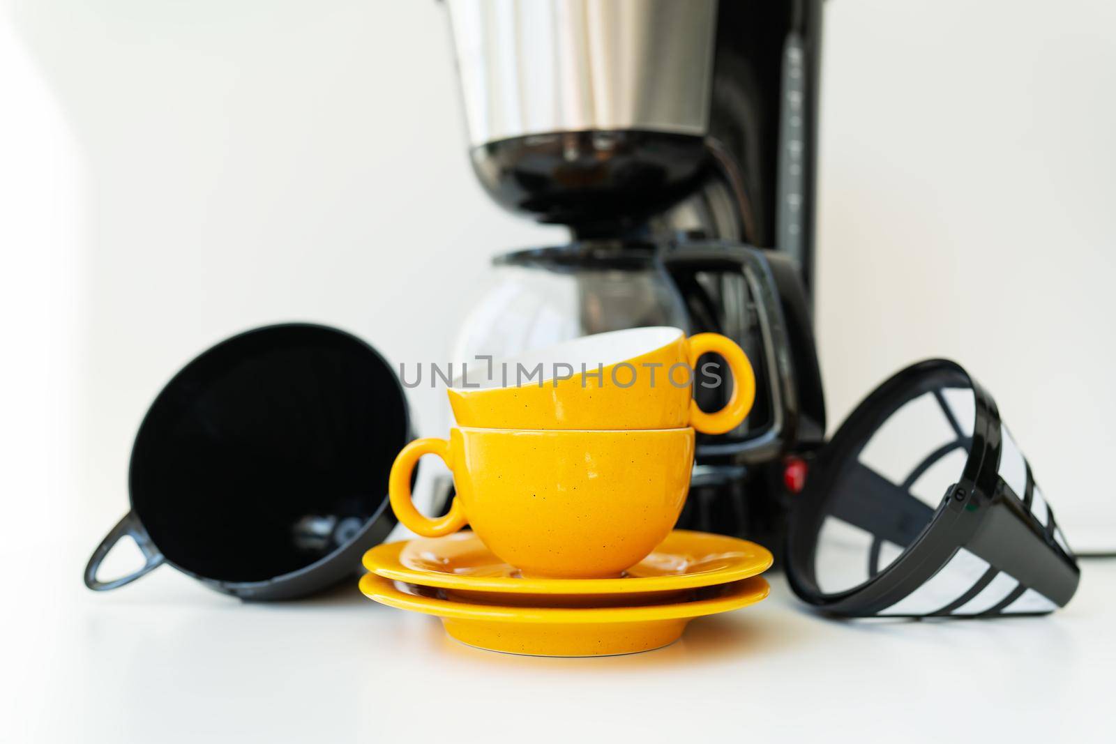 An automatic drip coffee maker stands with a yellow cup on a white table. Black modern drip coffee pot. Electric kitchen small household appliances. Appliances. by sfinks