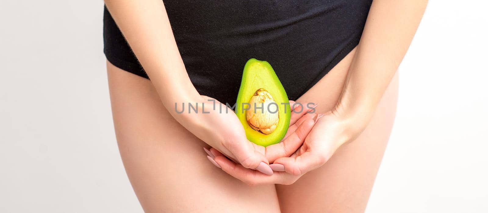 Young woman holding one half of a fresh avocado fruit close to her belly against a white background, healthy nutrition and pregnancy concept