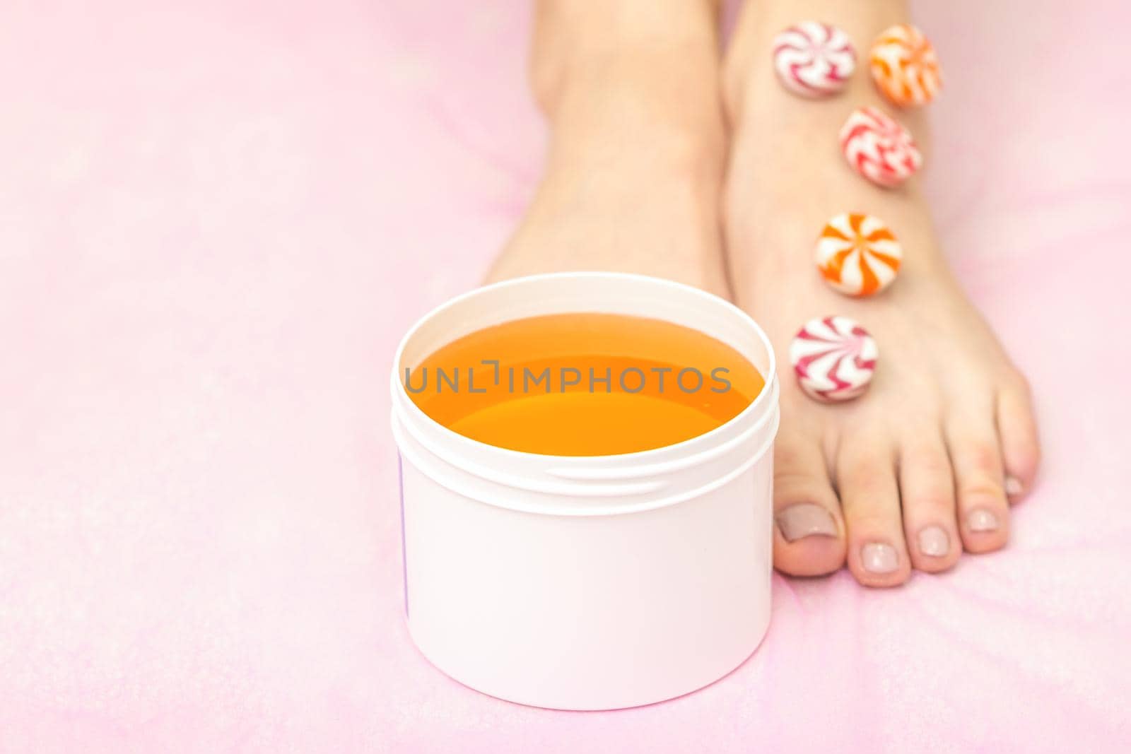 Round candies lying in a row on female feet with white jar with sugar paste on pink background with copy space, depilation concept
