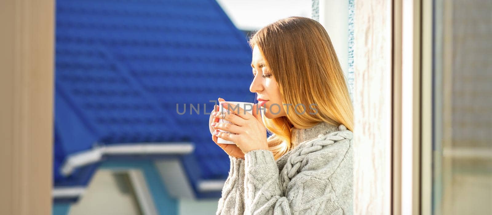 Woman relaxing on the balcony with coffee. A beautiful young woman in a sweater holds a cup of coffee standing near the window in the morning