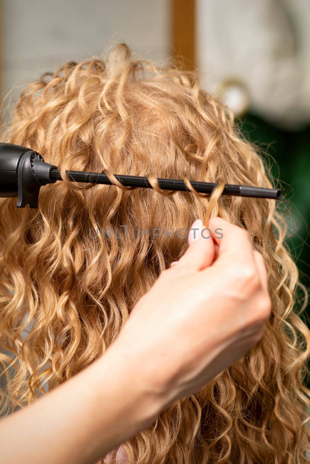 Hands of hairstylist curl wavy hair of young woman using a curling iron for hair curls in the beauty salon rear view. by okskukuruza