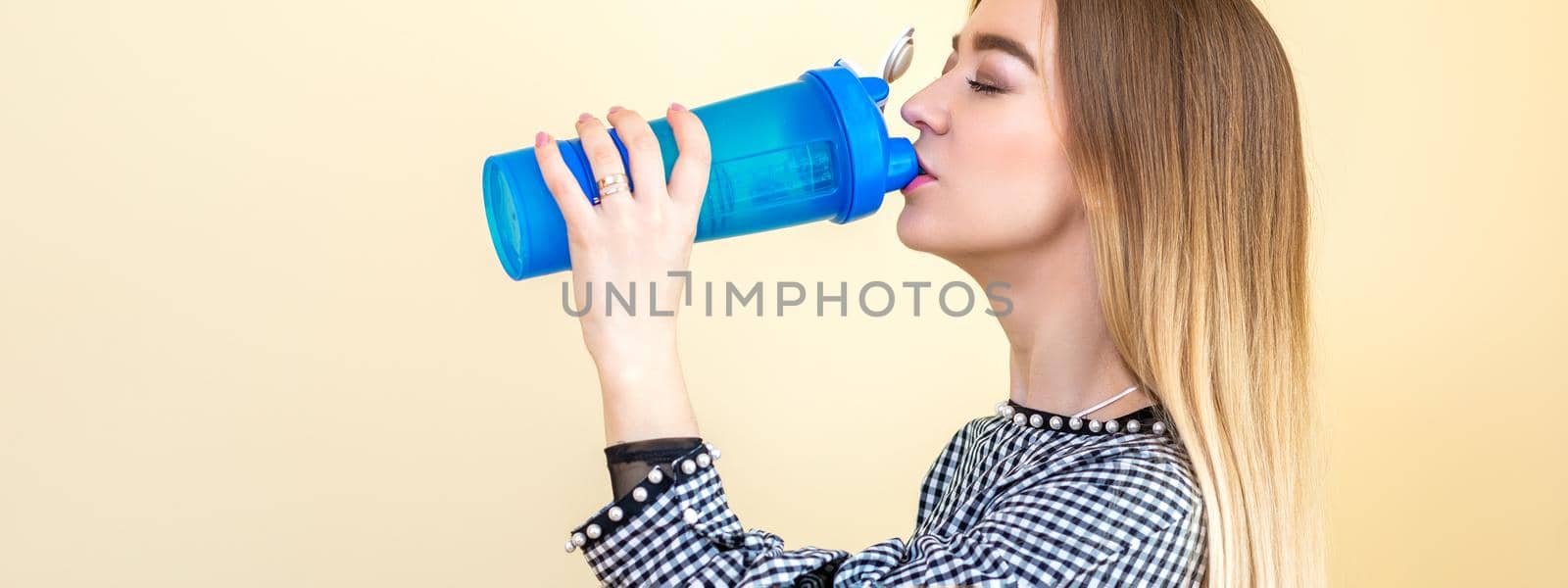 Thirsty woman with blue plastic bottle against a light background. A beautiful young caucasian businesswoman in a blouse drinking water on yellow