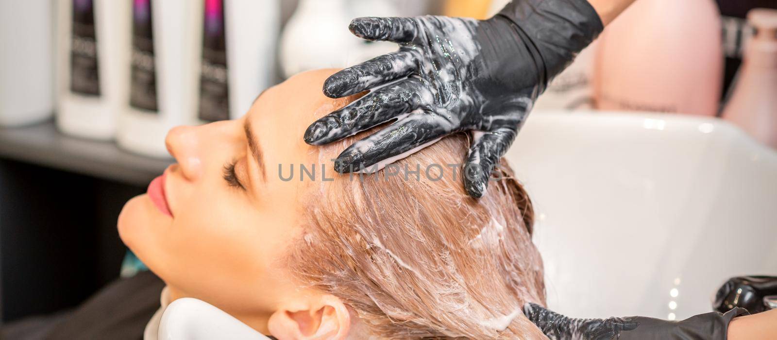 Young caucasian blonde woman having hair washed in the sink at a beauty salon. by okskukuruza