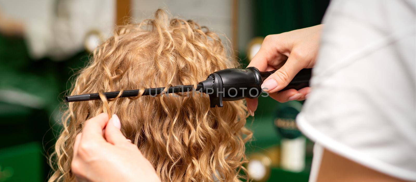 Hands of hairstylist curl wavy hair of young woman using a curling iron for hair curls in the beauty salon rear view