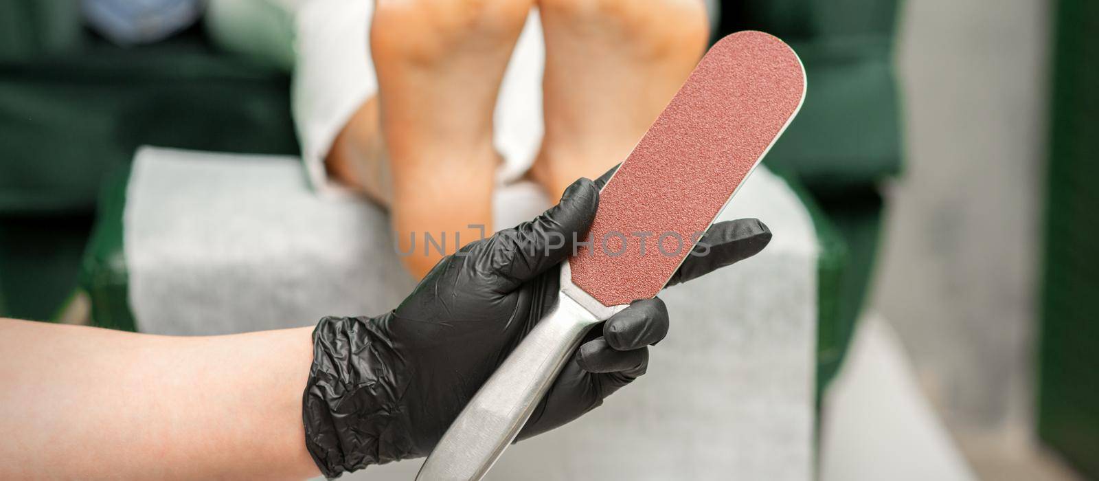 Special grater for scrubbing heels in hands of nail pedicure technician on feet background in beauty salon closeup. by okskukuruza