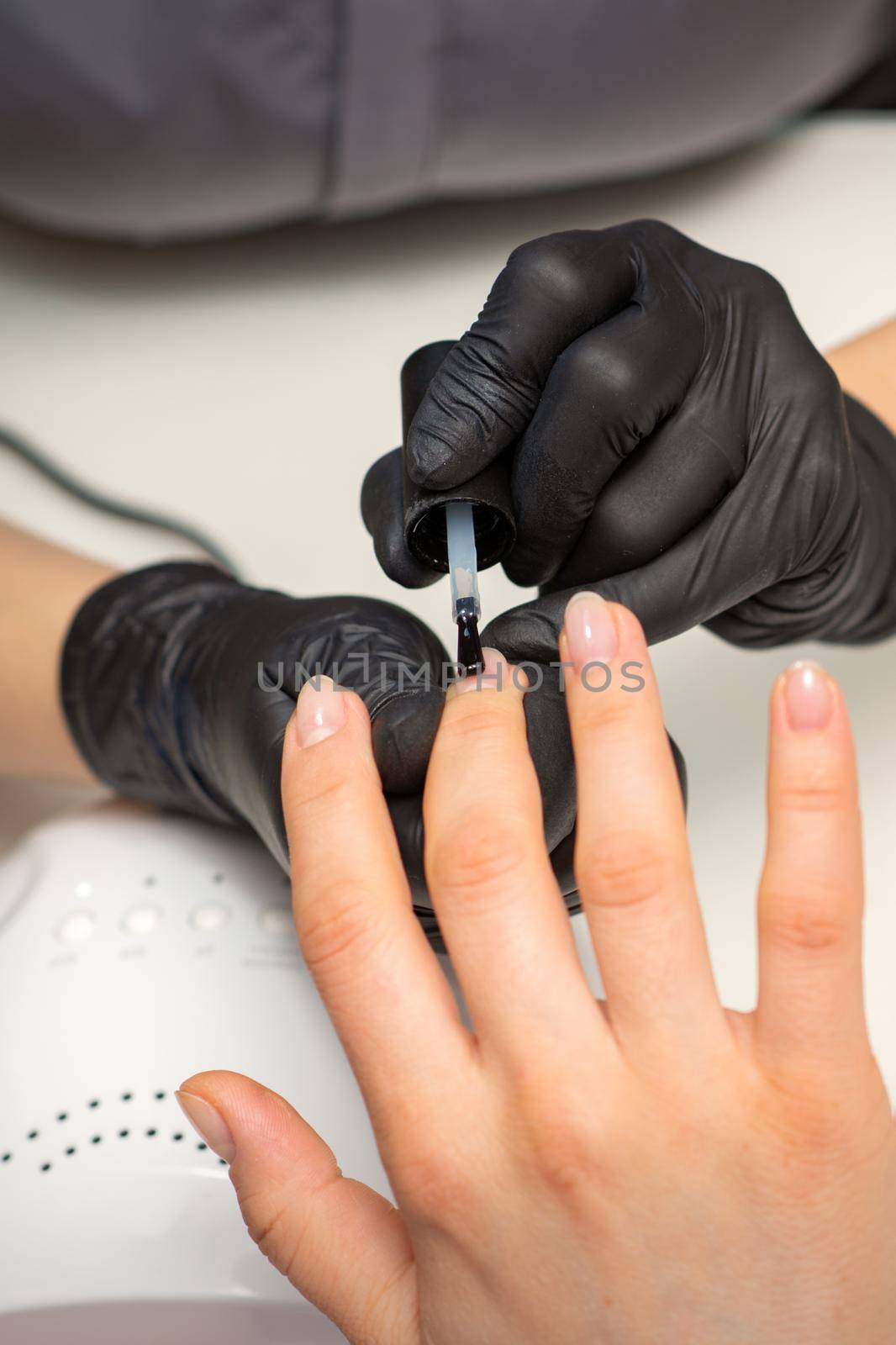 Painting nails of a woman. Hands of Manicurist in black gloves applying transparent nail polish on female Nails in a beauty salon