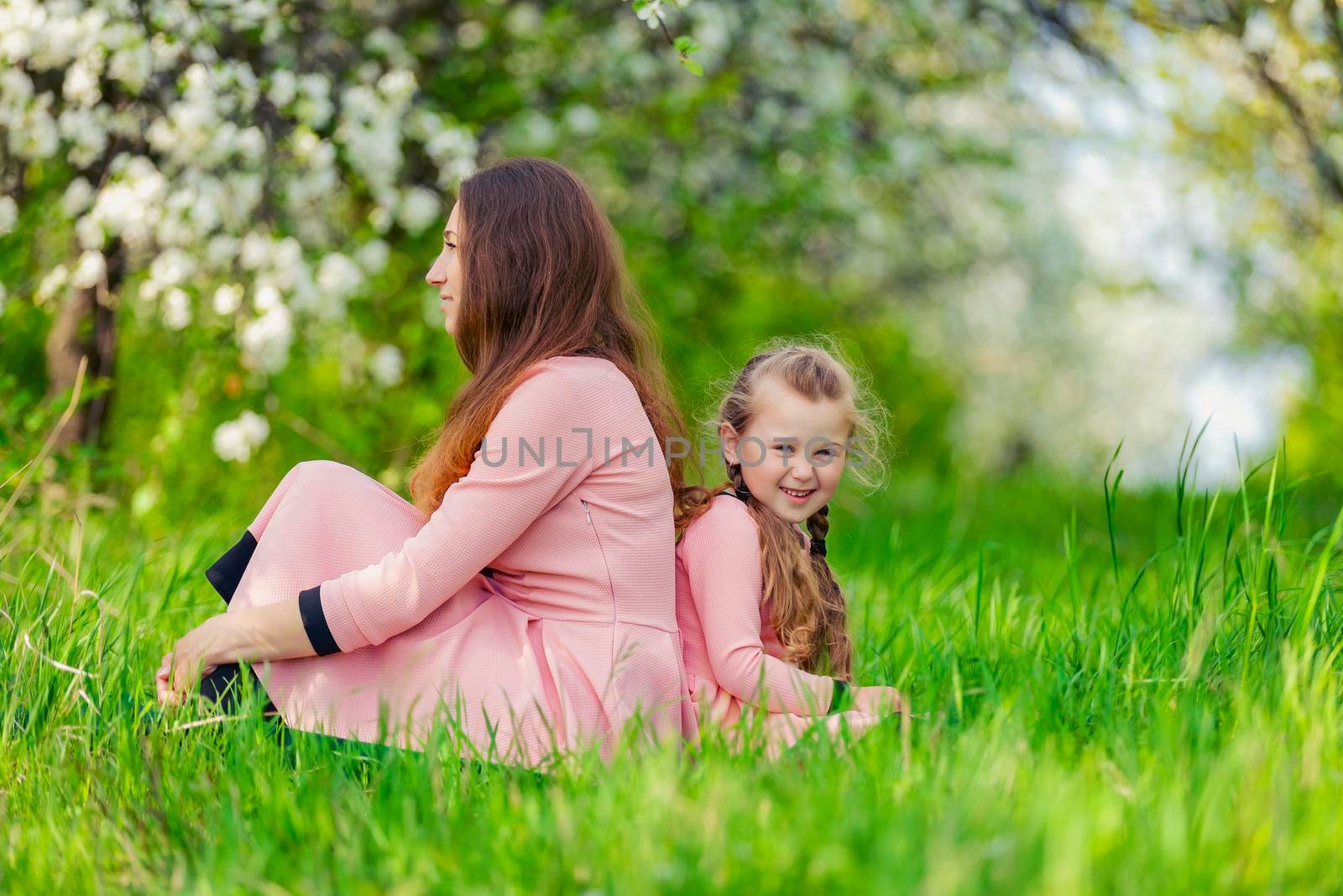 mother and daughter sit in a flowering garden with their backs to each other