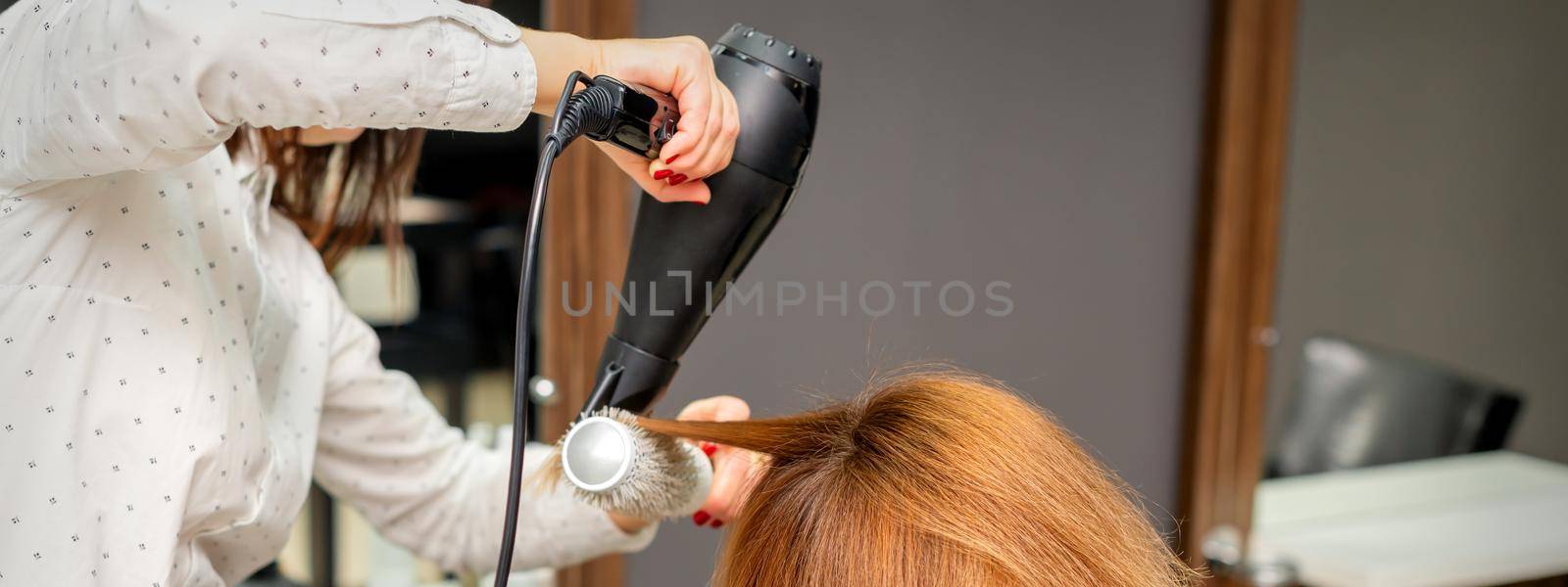 Drying hair in the hair studio. Female hairdresser stylist dries hair with a hairdryer and round brush red hair of a woman in a beauty salon. by okskukuruza