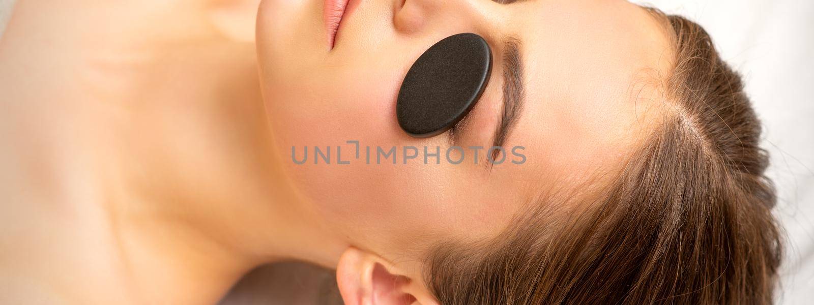 Black massage stones lying on the eyes of the young caucasian woman. Facial massage in a spa