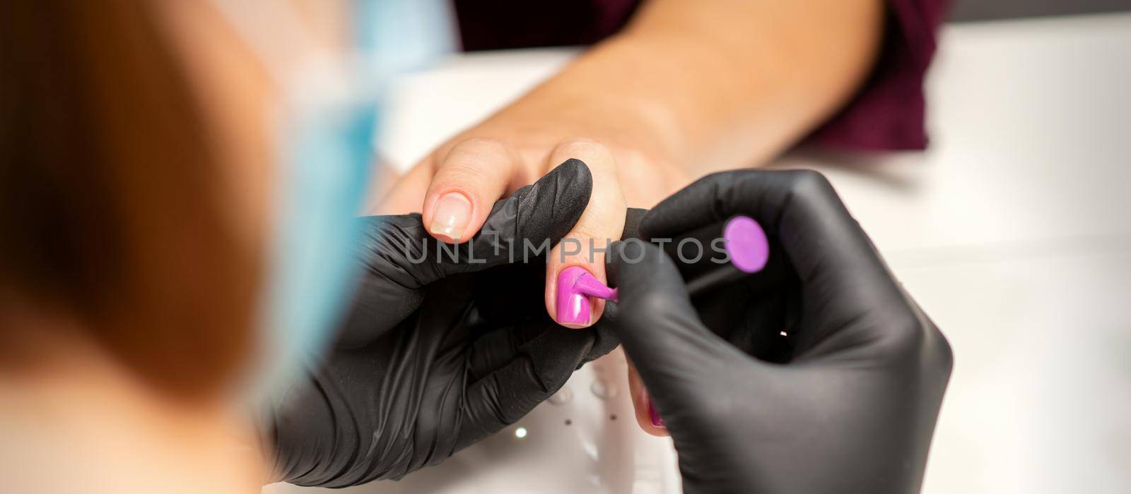 Painting nails of a woman. Hands of Manicurist in black gloves applying pink nail polish on female Nails in a beauty salon