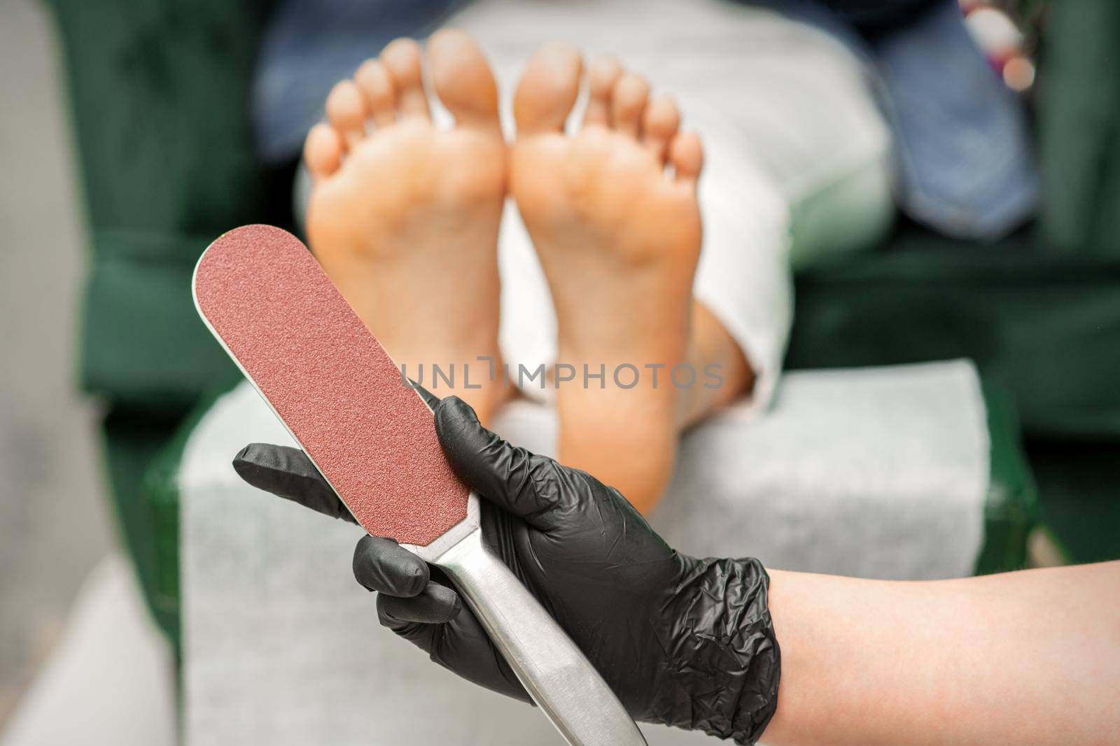 Special grater for scrubbing heels in hands of nail pedicure technician on feet background in beauty salon closeup. by okskukuruza