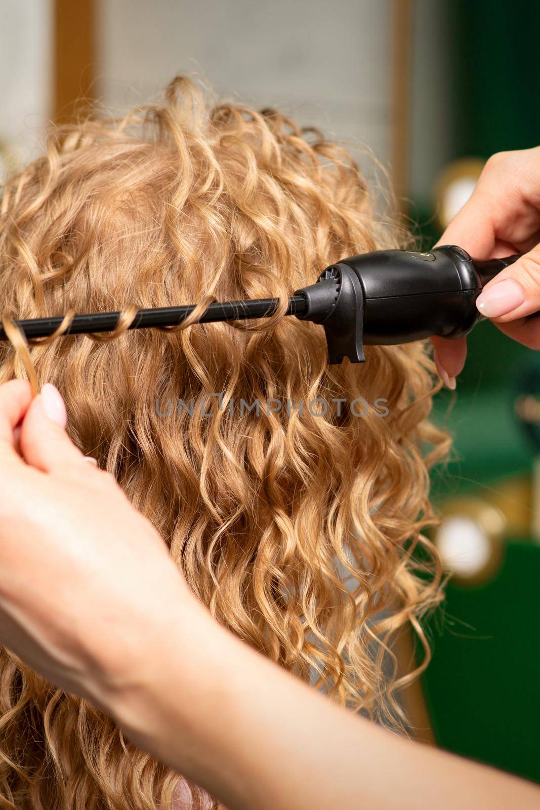 Hands of hairstylist curl wavy hair of young woman using a curling iron for hair curls in the beauty salon rear view