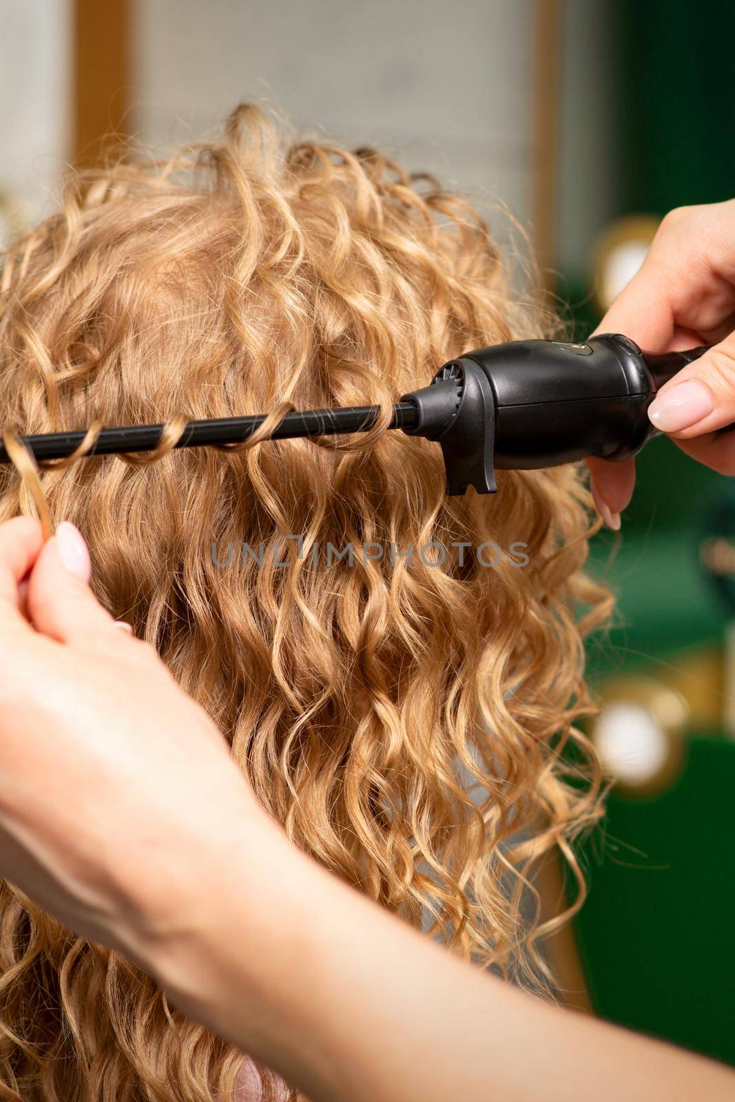 Hands of hairstylist curl wavy hair of young woman using a curling iron for hair curls in the beauty salon rear view. by okskukuruza