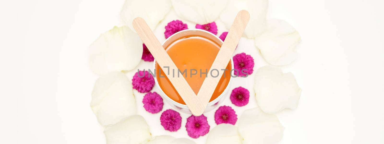 Jar of wax with wooden sticks, pink flowers, and white petals on white background. Concept of depilation. Flat lay top view. by okskukuruza