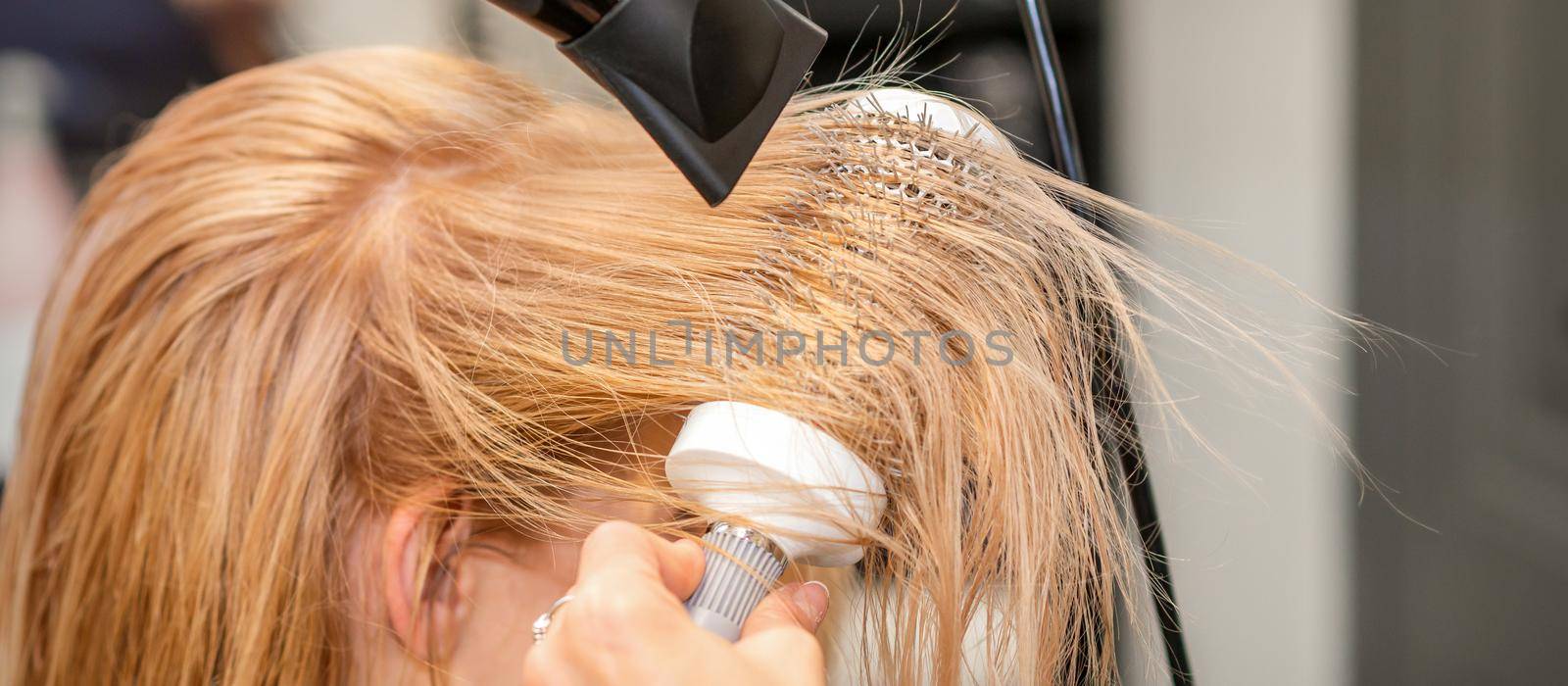 Hairdresser hand drying blond hair with a hairdryer and round brush in a beauty salon