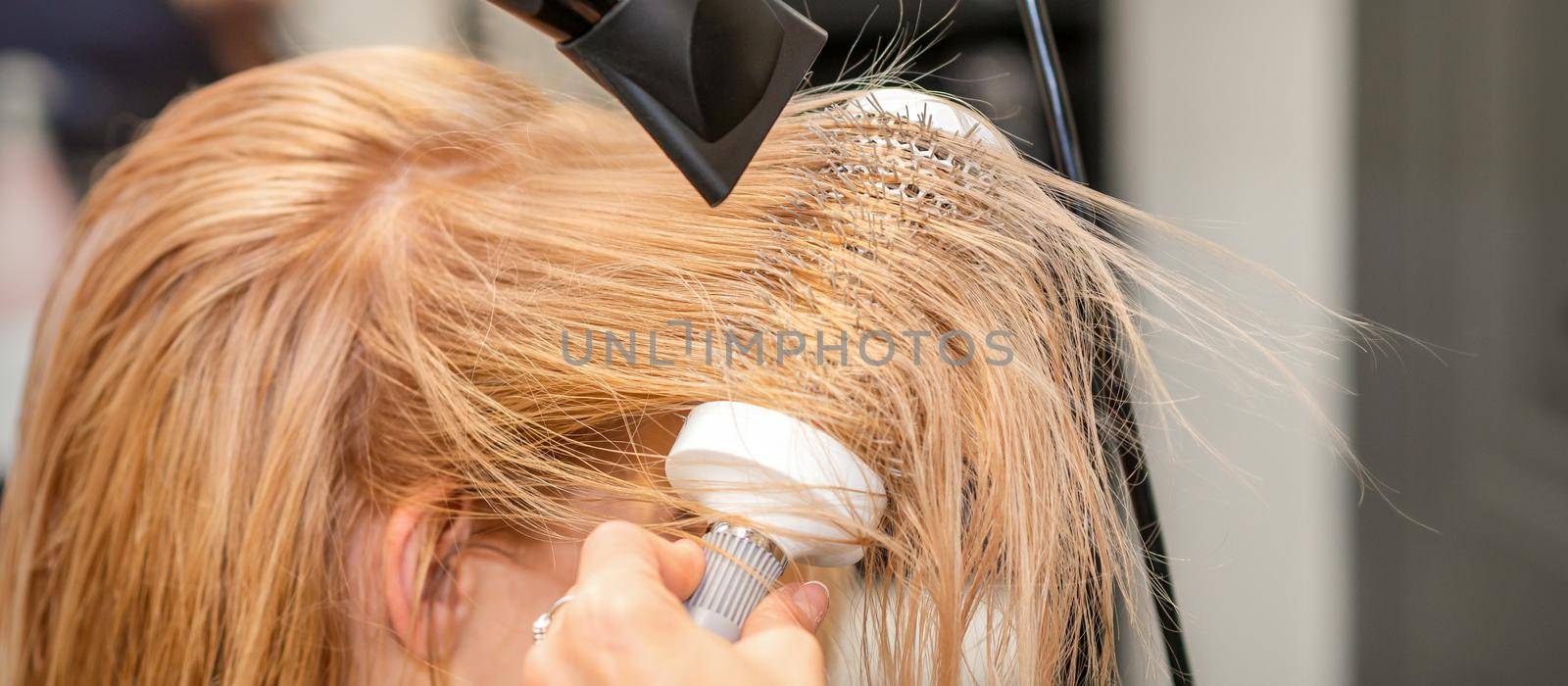 Hairdresser hand drying blond hair with a hairdryer and round brush in a beauty salon. by okskukuruza