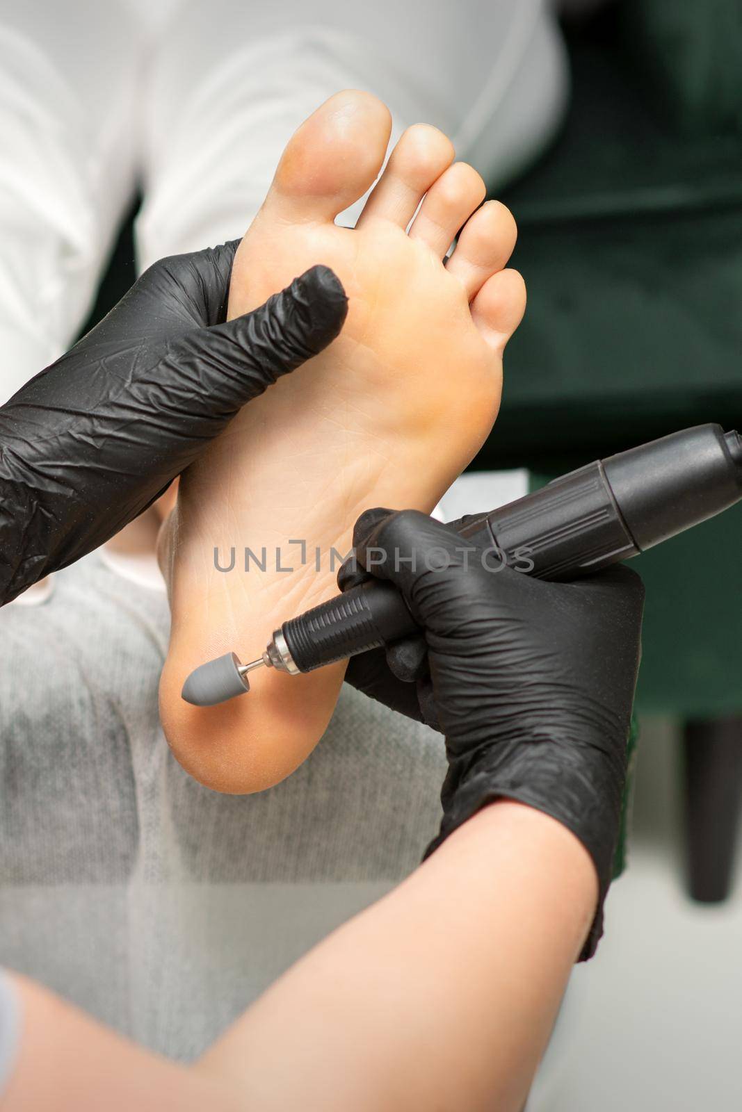 Peeling female feet. Hands of podiatry specialist in black gloves using special grinding equipment polish female foot from callus and corn in the beauty salon. by okskukuruza