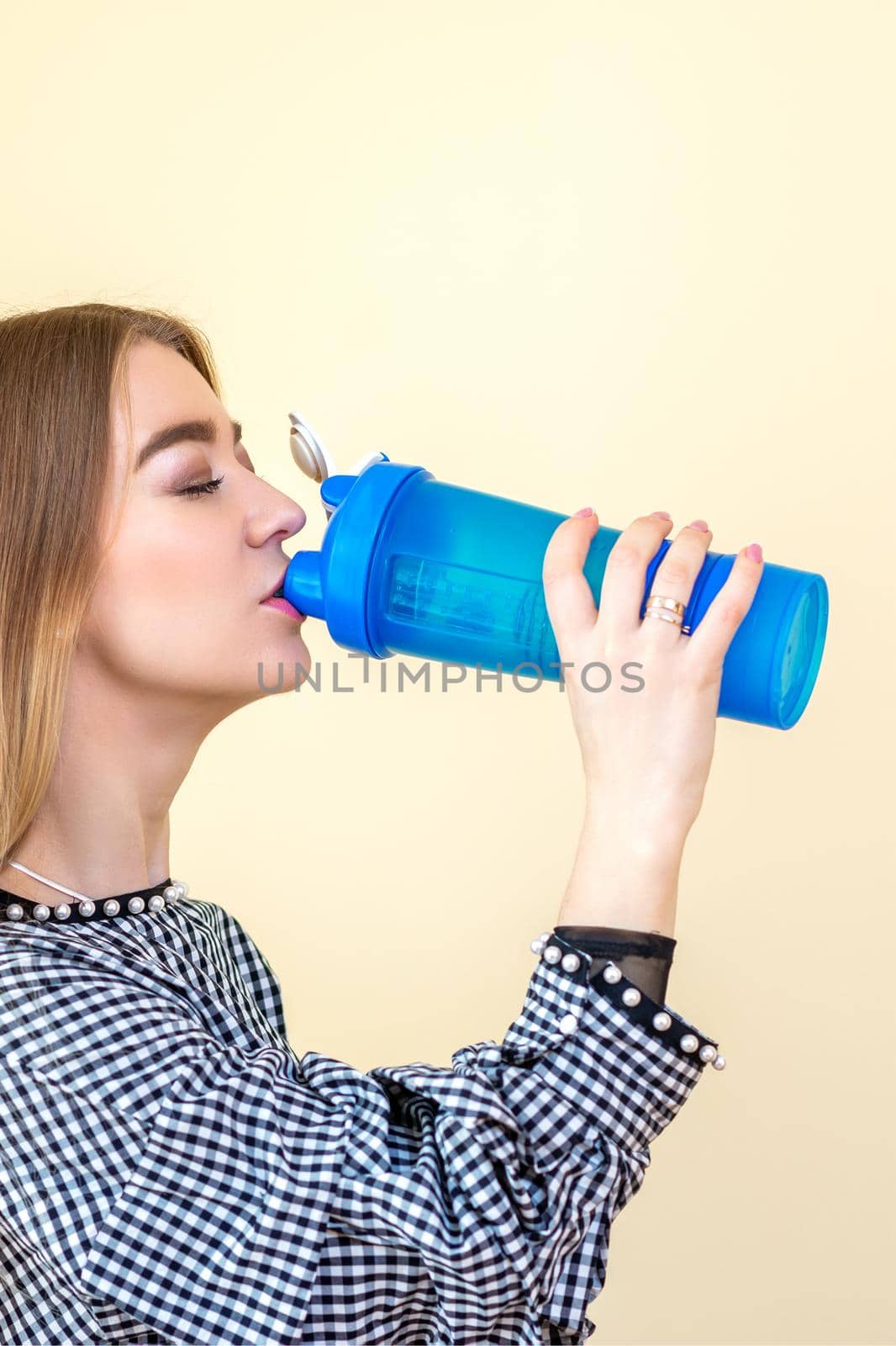 Thirsty woman with blue plastic bottle against a light background. A beautiful young caucasian businesswoman in a blouse drinking water on yellow