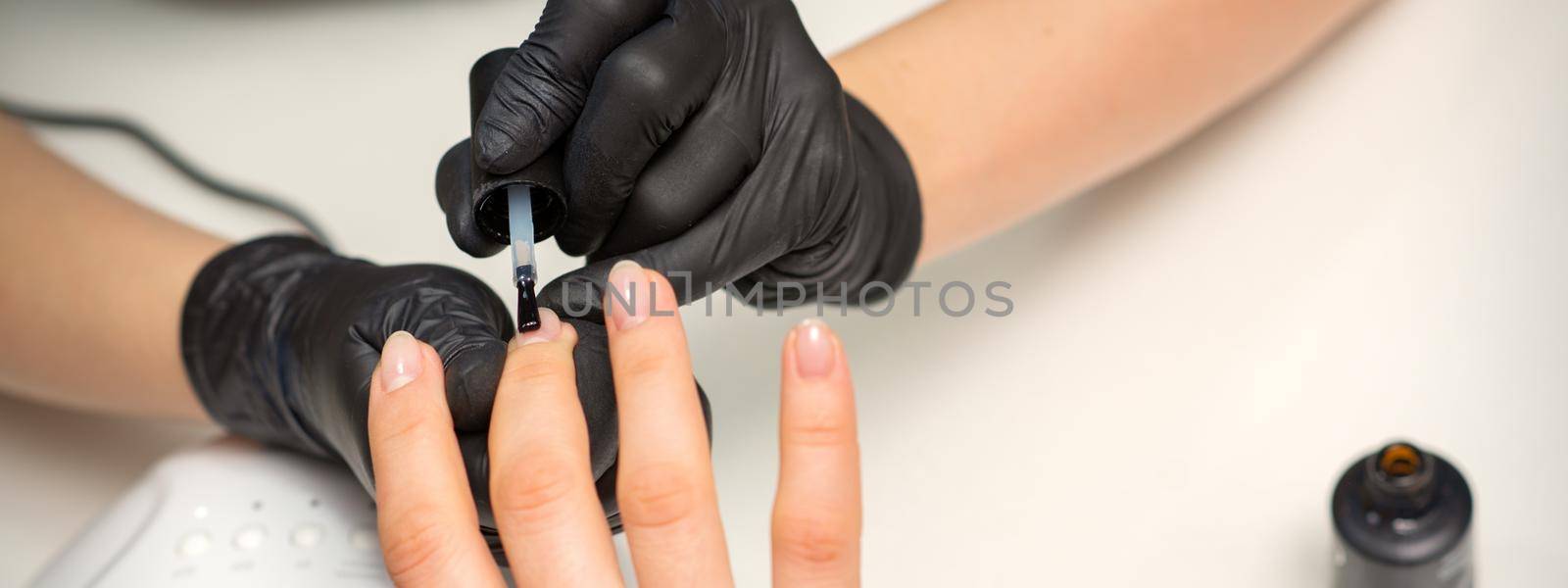 Painting nails of a woman. Hands of Manicurist in black gloves applying transparent nail polish on female Nails in a beauty salon. by okskukuruza