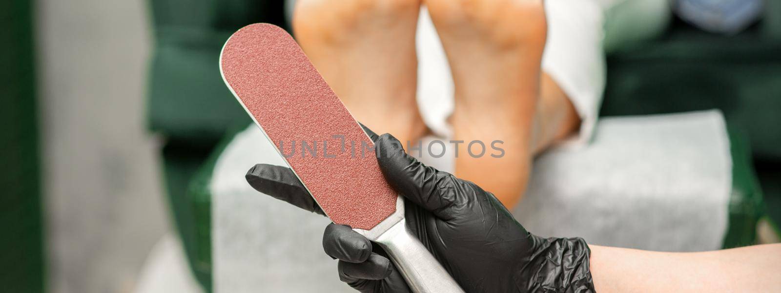 Special grater for scrubbing heels in hands of nail pedicure technician on feet background in beauty salon closeup