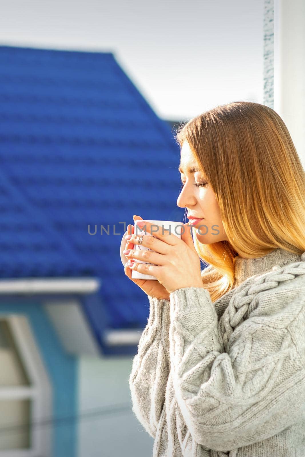 Woman relaxing on the balcony with coffee. A beautiful young woman in a sweater holds a cup of coffee standing near the window in the morning. by okskukuruza