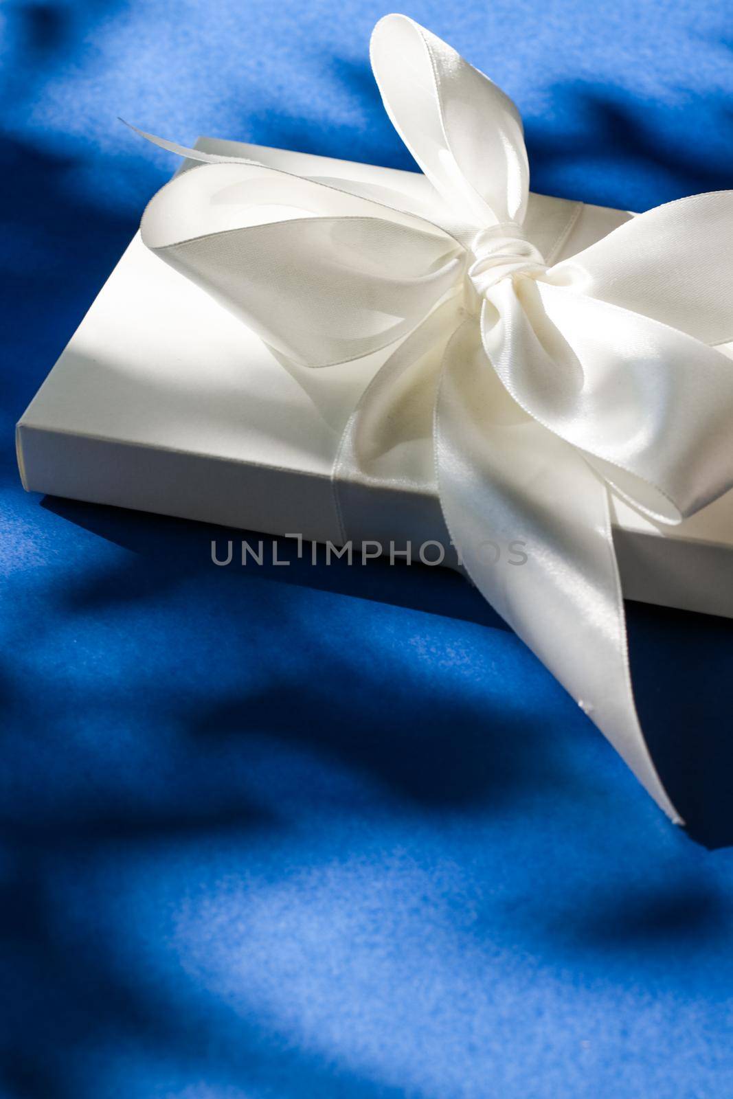 Luxury holiday white gift box with silk ribbon and bow on blue background, luxe wedding or birthday present by Anneleven