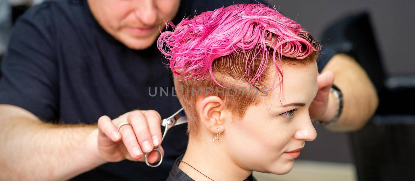Woman having a new haircut. A male hairstylist is cutting dyed pink short hair with scissors in a hair salon