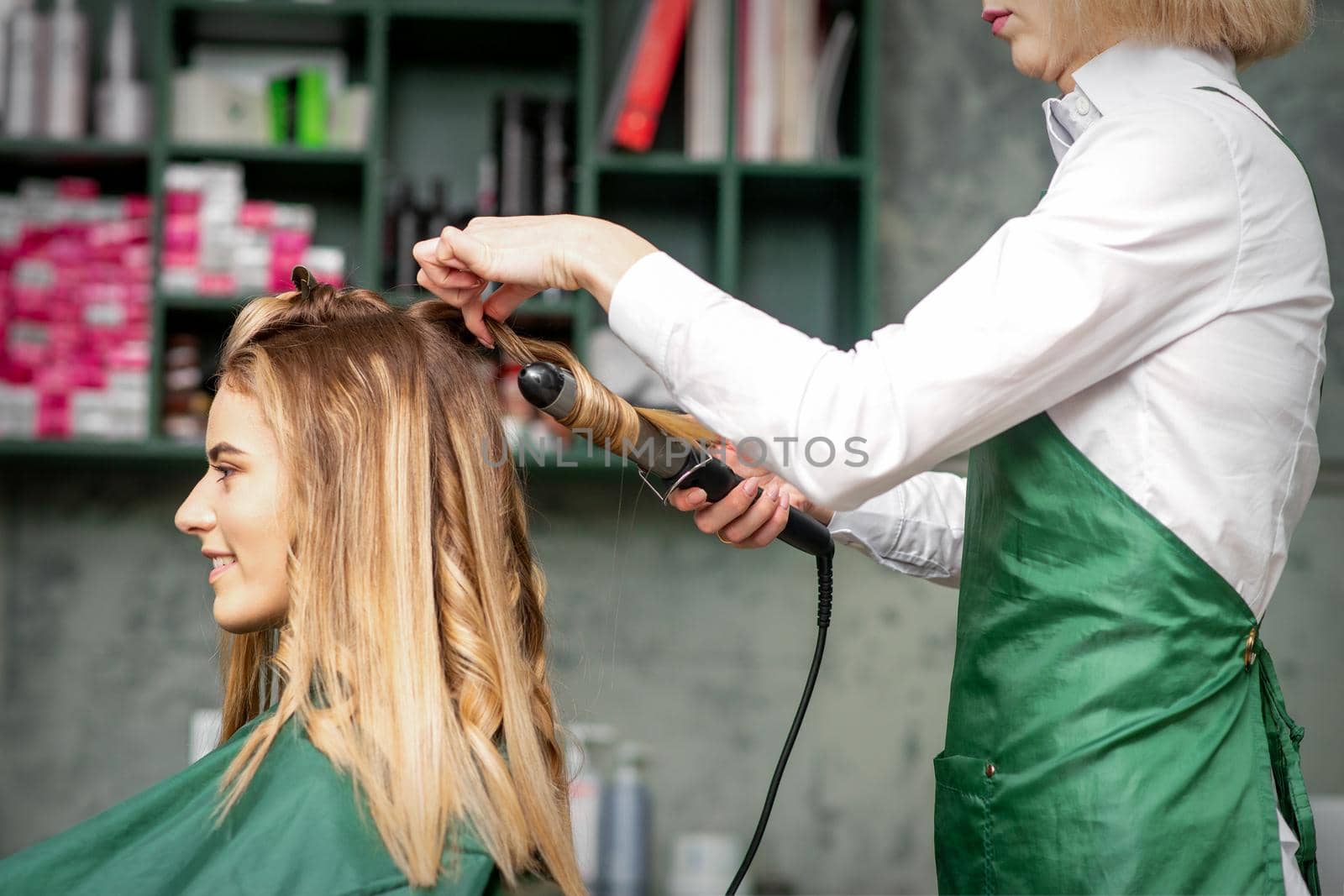 Creating curls with curling irons. Hairdresser makes a hairstyle for a young woman with long red hair in a beauty salon