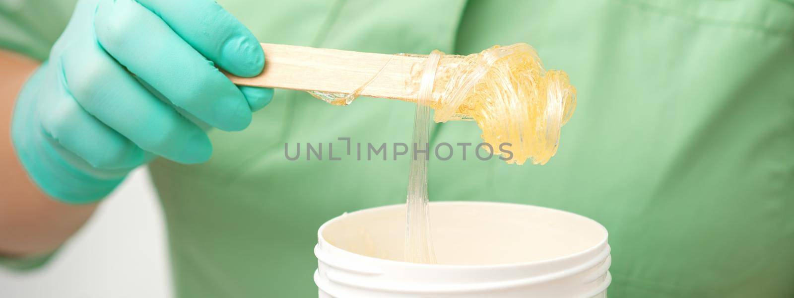 Liquid yellow sugar paste for depilation on a stick flows into the jar in hands of a beautician. by okskukuruza