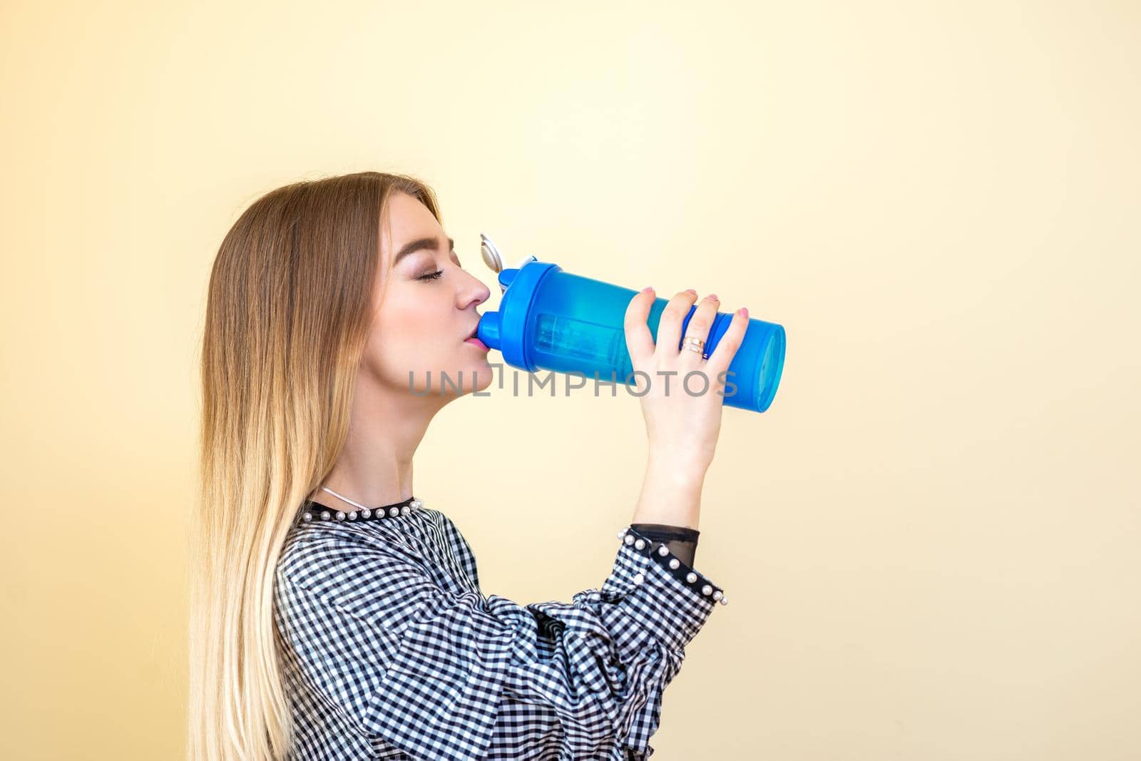 Thirsty woman with blue plastic bottle against a light background. A beautiful young caucasian businesswoman in a blouse drinking water on yellow