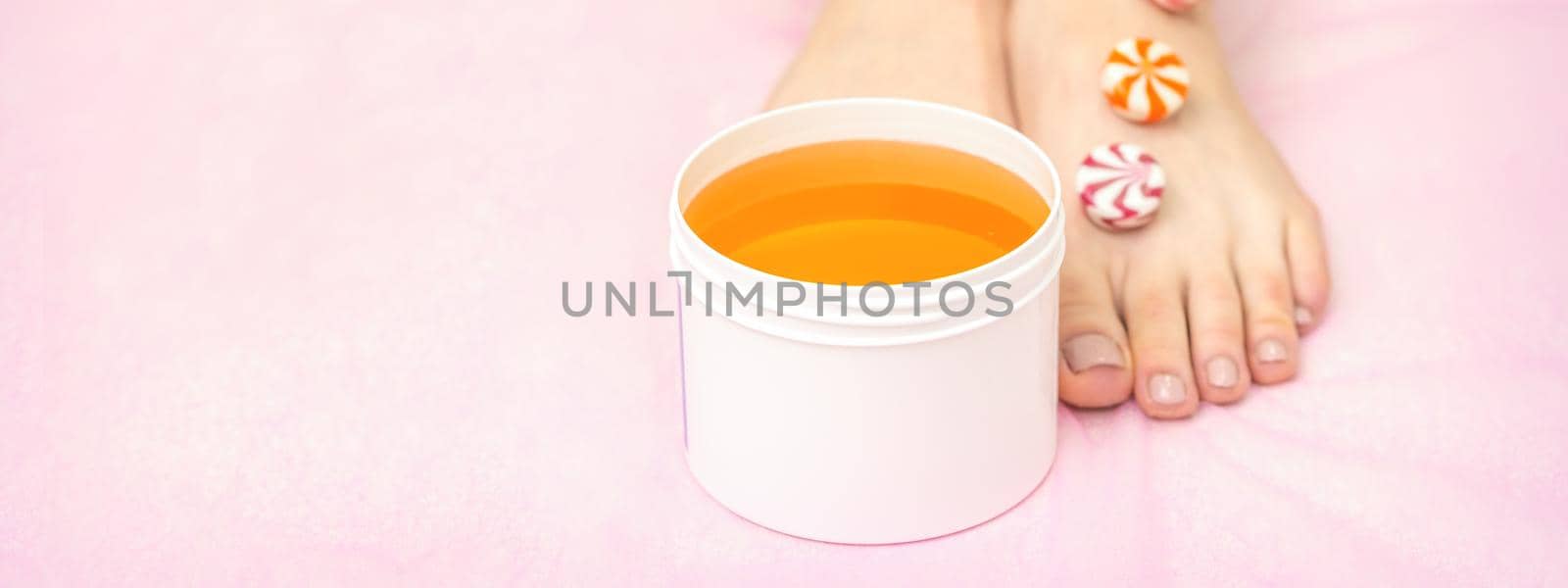 Round candies lying in a row on female feet with white jar with sugar paste on pink background with copy space, depilation concept