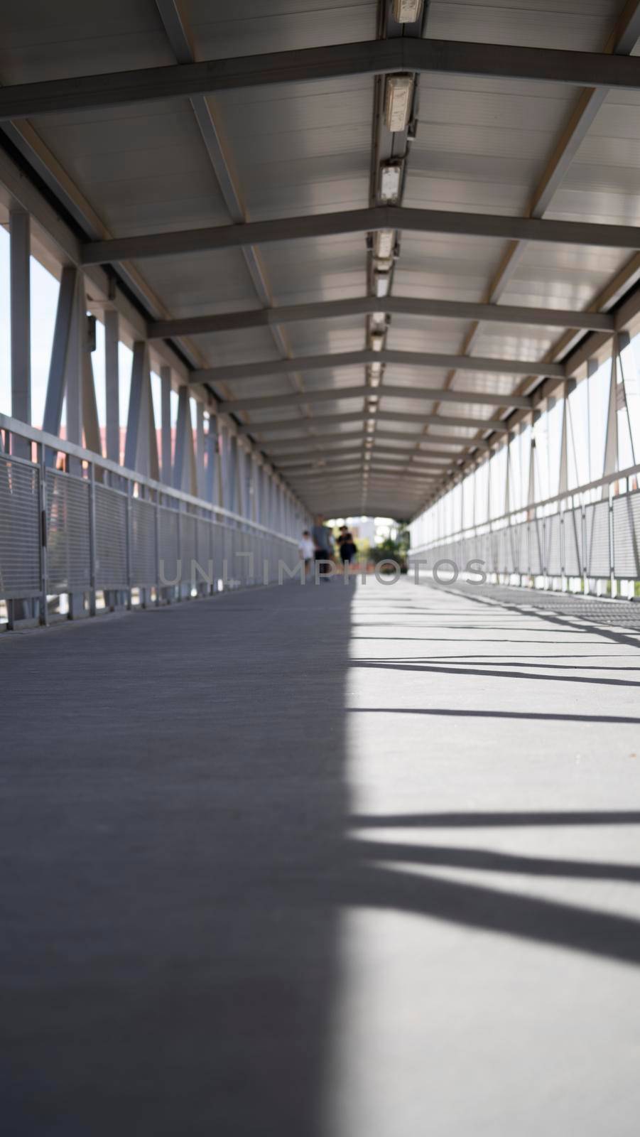 Pedestrian crosswalk under roof passage in urban environment. Overhead pedestrian bridge overpass in urban area. Footbridge tunnel with some pedestrians crossing through it.