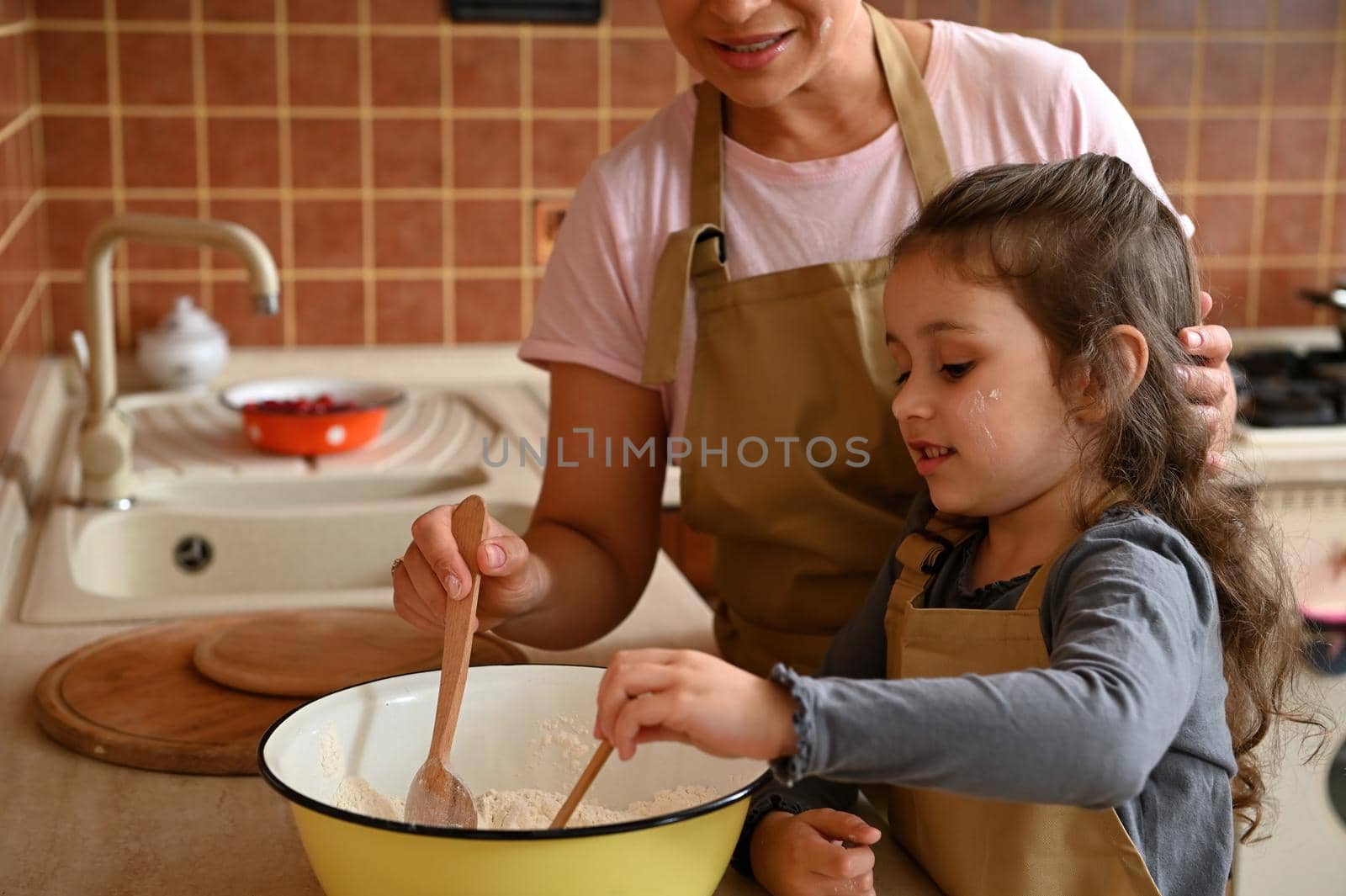 Caucasian cute little girl wearing a beige chef's apron, kneading a dough, standing near her mother in the home kitchen and learning preparing pie. Baking concept. Mom and daughter cooking together