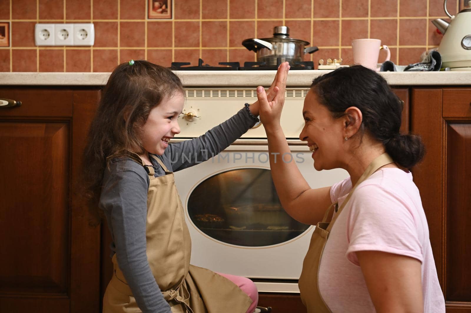 Loving mom in beige chef apron and her cute little daughter high five while standing by the oven in home kitchen by artgf