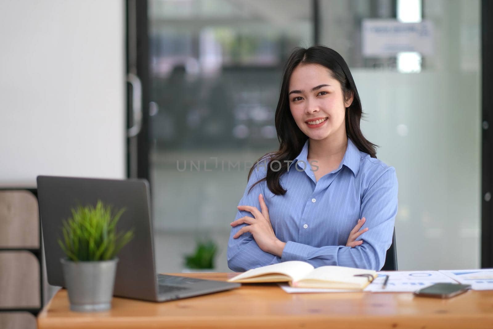 Confident young Asian business woman sitting with arms crossed smiling looking at camera in the office..