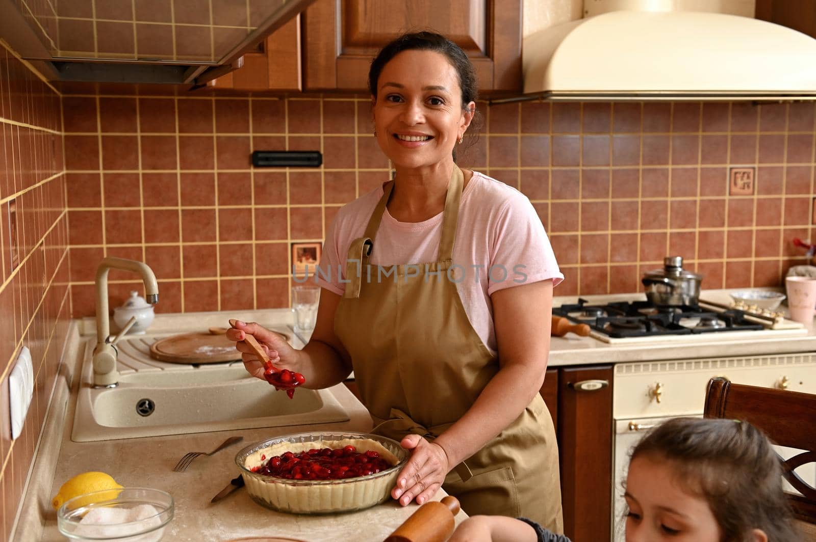 Beautiful middle-aged Latin American woman, loving mom in chef apron, putting fresh cherries on a rolled out dough, smiling a cheerful toothy smile while preparing homemade pie with her cute daughter