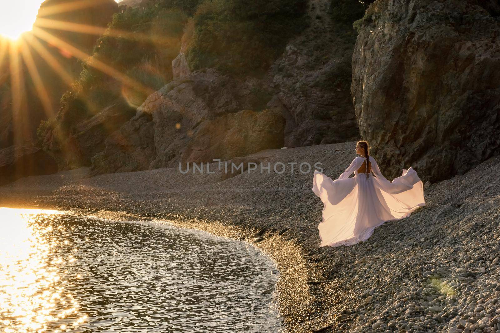 A mysterious female silhouette with long braids stands on the sea beach with mountain views, Sunset rays shine on a woman. Throws up a long white dress, a divine sunset. by Matiunina
