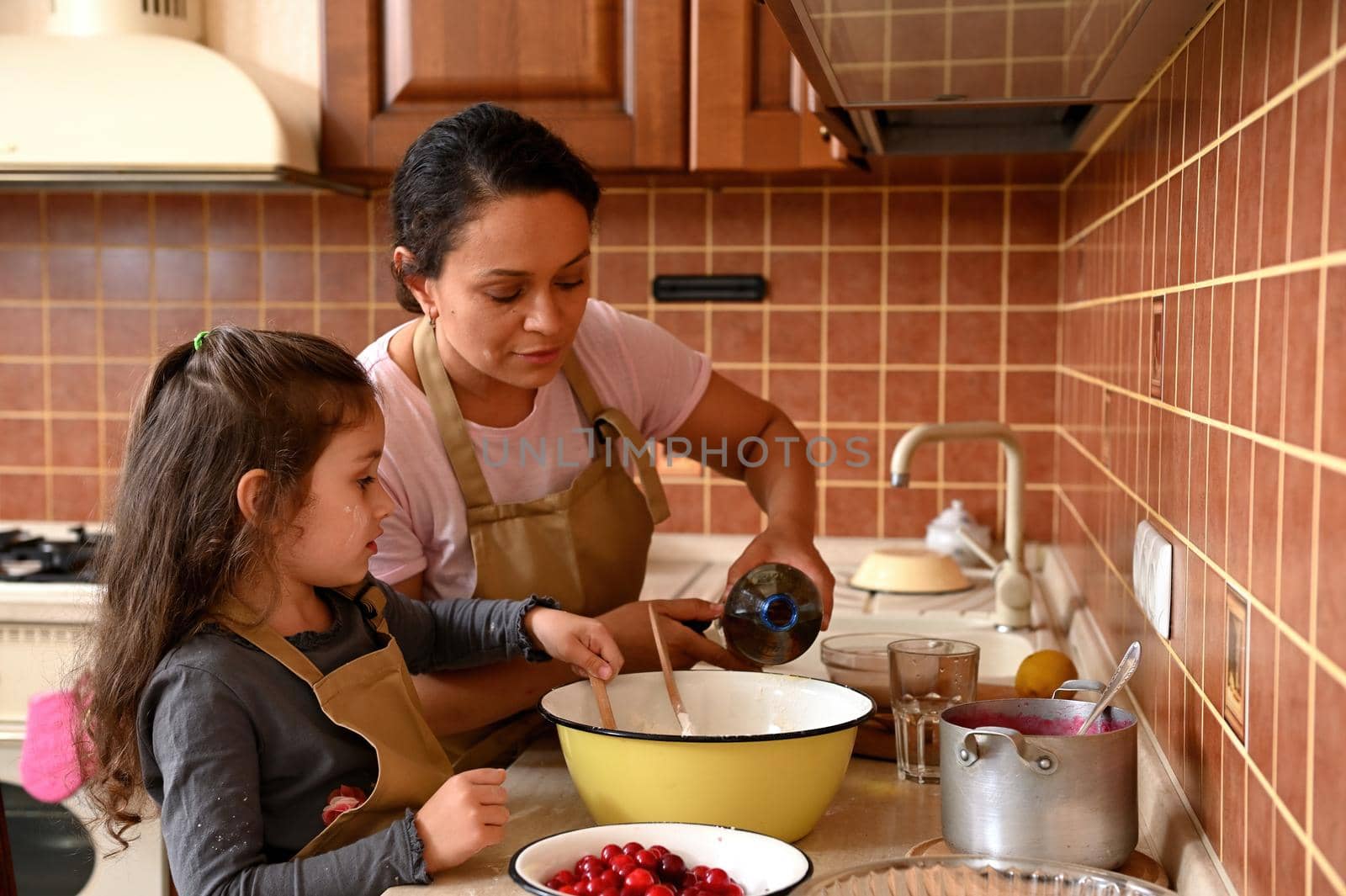 Mom and daughter cooking together, preparing dough for homemade festive cherry pie. Charming woman pouring some oil into a vintage enamel bowl, adding ingredients. Baking concept. Family relationships